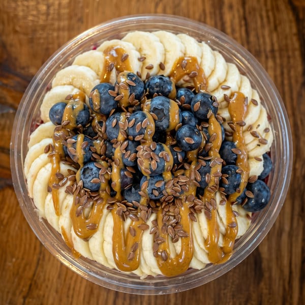 Overhead view of a fruit bowl with sliced bananas, blueberries, peanut butter drizzle, and flax seeds.