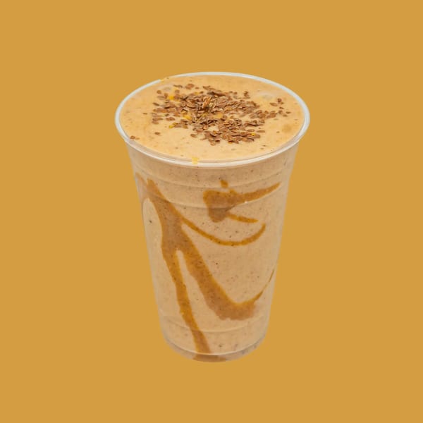 A light brown smoothie with a caramel swirl and flax seeds on top, in a clear plastic cup.